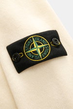 Close-up angled view of the Stone Island Crew Neck Sweater 'Lambswool' creme showing the iconic black compass badge with green and yellow embroidery on a warm, soft virgin wool and microfiber knit. Stylish essential for the cold season with distinctive li