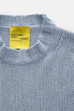 Close-up front view of B.F. Knitwear Cashmere crew neck sweater light blue melange showing medium coarse knit, elastic ribbed cuffs, and label. Autumnal cashmere from the knitting expert B.F. KNITWEAR: This softly knitted crew neck sweater is the perfect 
