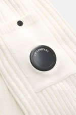 Nahaufnahme des kremfarbenen C.P. Company Rundhalspullovers mit Rippstrick aus reiner Baumwolle, weicher Qualität, mittelfeinem Strickbild, Slim Fit, Raglanärmel, Tasche am Ärmel und labeltypischem Linsen-Detail, schräg von oben fotografiert.