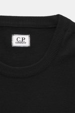 Nahaufnahme des C.P. Company Rundhalspullover schwarz, Frontansicht mit Rundhalsausschnitt, feinem Strickbild, weichem Griff, Slim Fit, Tasche am Oberarm, flexible Rippbündchen, Mix aus Schurwolle und Microfaser, labeltypisches Linsen-Detail.