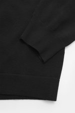 Nahaufnahme des C.P. Company Rundhalspullover schwarz von schräg oben, zeigt weichen Griff, feines Strickbild, Slim Fit, Rundhalsausschnitt, Tasche mit Druckknopf am Oberarm, labeltypisches Linsen-Detail und flexible Rippbündchen, Mix aus Schurwolle und M