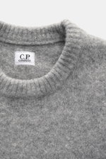 Nahaufnahme aus Draufsicht des C.P. Company Rundhalspullover grau meliert, mit mittelfeinem Strickbild, weichem Griff, Slim Fit, elastischer Qualität, Zipper-Tasche am Oberarm, flexible Rippbündchen, Linsen-Detail, Mix aus Wolle und Microfaser.
