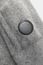 Nahaufnahme des C.P. Company Rundhalspullover grau meliert von schräg oben, zeigt weichen, voluminösen Griff, mittelfeines Strickbild, elastische Qualität, Slim Fit, Zipper-Tasche am Oberarm, labeltypisches Linsen-Detail, flexible Rippbündchen, Mix aus Wo