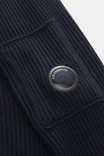 Nahaufnahme des C.P. Company Rundhalspullover navy von schräg oben, mittelfeines Strickbild, weicher Griff, Patent-Strick, Slim Fit, Tasche mit Zipper am Oberarm, labeltypisches Linsen-Detail, flexible Rippbündchen, Strick aus Schurwolle und Microfaser.