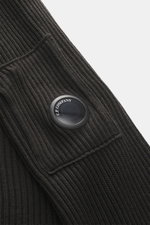 Nahaufnahme des C.P. Company Rundhalspullover anthrazit, schräg fotografiert, mittelfeines Strickbild, weicher Griff, Slim Fit, Rundhalsausschnitt, Tasche mit Zipper am Oberarm, labeltypisches Linsen-Detail, flexible Rippbündchen, Strick aus Schurwolle un