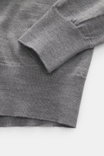 Nahaufnahme des Ärmels und Saums eines Ami Paris Merino Strickpolo grau meliert, reine Merinowolle, weicher Griff, feines Strickbild, Regular Fit, typischer Polokragen, Drei-Knopf-Leiste, elastische Rippbündchen, Logo-Stitching, fotografiert von oben.