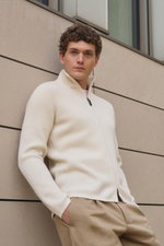 Junger Mann lehnt seitlich an Wand, trägt von Braun Cashmere Strickjacke offwhite mit hohem Kragen, Slim Fit, Patentstrick und Zwei-Wege-Reißverschluss.