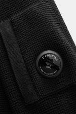 Nahaufnahme der schwarzen C.P. Company Strickjacke mit puristischem Design, Waffelstrick-Optik und labeltypischem NFC-Linsen-Detail am Oberarm.