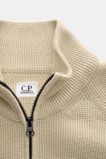 Nahaufnahme der C.P. Company Strickjacke sand im puristischen Design aus mittelfeinem Baumwoll-Microfaser-Strick mit Waffelstruktur, Stehkragen und robustem Zwei-Wege-Reißverschluss, fotografiert aus leicht seitlicher Vogelperspektive.