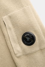 Nahaufnahme der C.P. Company Strickjacke sand mit markanter Waffelstrick-Optik, Zipper-Tasche am Oberarm und auffälligem Linsen-Detail.