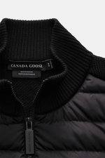 Nahaufnahme aus schräger Perspektive der Canada Goose Strickjacke 'Hybridge Knit' schwarz mit hochschließendem Stehkragen aus reiner Wolle und Daunen-Front, robuster Reißverschluss sichtbar.