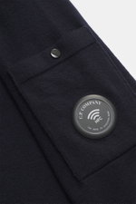 Nahaufnahme der C.P. Company Strickjacke navy am Oberarm, Strick aus Schurwolle, feines Strickbild, Regular Fit, Zwei-Wege-Reißverschluss, Stehkragen, Tasche mit Knopf, labeltypisches Linsen-Detail sichtbar.