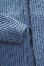 Nahaufnahme der graublauen C.P. Company Strickjacke, Perspektive zeigt voluminöses Strickbild, robusten Zwei-Wege-Reißverschluss und weiche Qualität aus Schurwolle, Slim Fit mit gestrickter Kapuze, schräge Eingriffstaschen, Zipper-Tasche am Oberarm und la