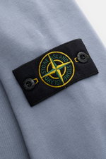 Alt tag (125 characters max):
Close-up side view of Stone Island Rundhals-Sweatshirt 'Basic Sweat' rauchblau showing black compass badge on soft cotton fabric.

Additional description:
<p>Zeitloses Basic Piece: Das sportive Sweatshirt von STONE ISLAND wir