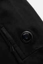 Nahaufnahme des schwarzen C.P. Company Sweatshirts aus reiner Baumwolle, zeigt die Ärmel-Tasche mit ikonischem Linsen-Detail, fotografiert aus seitlicher Perspektive.