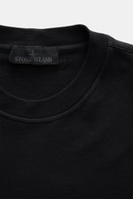 Nahaufnahme des schwarzen Stone Island Sweatshirts 'Ghost Piece' mit Rundhalsausschnitt, weichem Griff, elastischer Baumwollqualität und tonalem Kompass-Badge am Ärmel, fotografiert von oben.