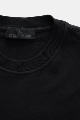 Nahaufnahme des schwarzen Stone Island Sweatshirts 'Ghost Piece' mit Rundhalsausschnitt, weichem Griff, elastischer Baumwollqualität und tonalem Kompass-Badge am Ärmel, fotografiert von oben.