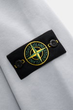 Close-up top-down view of the Stone Island Rundhals-Sweatshirt 'Basic Sweat' rauchblau showing the black compass badge with yellow and green embroidery on soft light blue cotton fabric.

<p>Zeitloses Basic Piece: Das sportive Sweatshirt von STONE ISLAND w