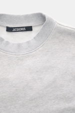 Nahaufnahme des Rundhalsausschnitts eines Jacquemus Sweatshirts 'Torneo' hellgrau meliert, Sweat aus reiner Baumwolle, softe Innenseite, Regular Fit, Patch am Saum, Logo-Stitching auf der Rückseite, komfortable Rippbündchen, fotografiert von oben.