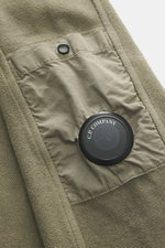 Nahaufnahme des C.P. Company Sweat-Troyer taupe von schräg oben, zeigt aufgesetzte Tasche am Oberarm mit labeltypischem Linsen-Detail, Sweat aus reiner Baumwolle, weicher Griff, Regular Fit, Troyer-Kragen mit Reißverschluss und flexible Rippbündchen.