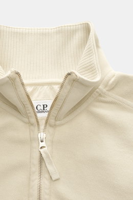 Nahaufnahme des C.P. Company Sweat-Troyer sand von schräg oben, Sweat aus reiner Baumwolle, weicher Griff, Regular Fit, Troyer-Kragen mit Reißverschluss, aufgesetzte Tasche am Oberarm, labeltypisches Linsen-Detail, flexible Rippbündchen.