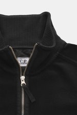 Nahaufnahme des schwarzen C.P. Company Sweat-Troyer von oben, Sweat aus reiner Baumwolle, weicher Griff, Regular Fit, Troyer-Kragen mit Reißverschluss, aufgesetzte Tasche am Oberarm, labeltypisches Linsen-Detail, flexible Rippbündchen.