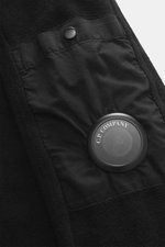 Nahaufnahme des schwarzen C.P. Company Sweat-Troyers aus reiner Baumwolle mit weichem Griff, Regular Fit, Troyer-Kragen, Reißverschluss, aufgesetzter Tasche am Oberarm, labeltypischem Linsen-Detail und flexiblen Rippbündchen, fotografiert von schräg oben.