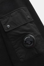 Nahaufnahme des schwarzen C.P. Company Sweatshirts aus reiner Baumwolle, Fokus auf Ärmeltasche mit Linsen-Detail, von oben fotografiert.