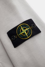 Stone Island Sweatjacke hellgrau, Detailaufnahme des typischen Kompass-Badges auf weichem, stückgefärbtem Baumwoll-Sweat aus schräger Perspektive.