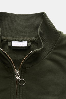 Nahaufnahme der dunkelgrünen 04651/ A trip in a bag Sweatjacke 'IF Track' aus Baumwoll-Jersey mit Reißverschluss, hoch geschlossenem Kragen und kleinem Weblabel, fotografiert von oben.
