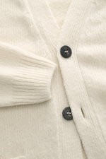 Nahaufnahme des cremefarbenen 04651/ A trip in a bag Cardigan 'Hunter' mit V-Ausschnitt, Vier-Knopf-Leiste, grobem Strickbild, weichem Griff, aufgesetzten Fronttaschen und Rippbündchen, fotografiert von oben.