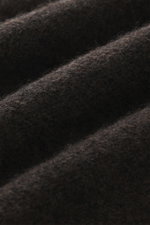 Nahaufnahme von dunkelbraun meliertem Strickstoff des 04651/ A trip in a bag Troyer 'Cloudy Wool' mit weichem Griff, mittelgrobem Strickbild, Regular Fit, Troyer-Kragen mit Knöpfen und breiten Rippbündchen, fotografiert aus seitlicher Perspektive.