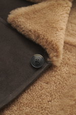 Nahaufnahme der Closed Lammfelljacke dunkelbraun von schräg oben, zeigt flauschiges Lammfell-Innenfutter, geschmeidiges Lammleder und Knopf.