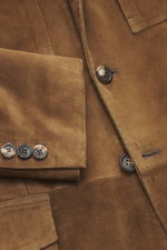 Close-up view of the Rifugio Brown Suede Blazer showing supple suede leather, soft texture, sleeve cuffs with buttons, patch chest pocket with flap, and three-button style. RIFUGIO combines style with safari elements in this luxurious jacket made of suppl