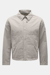 Stone Island Jacke 'Ghost Piece' hellgrau, frontal fotografiert, Slim Fit, Microfaser, Druckknopfleiste, Umlegekragen, Fronttaschen, Kompass-Badge, Ärmel mit Druckknopf.