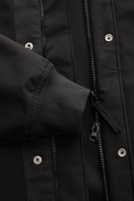 Close-up front view of Stone Island Blouson 'Light Soft Shell-R' black showing black three-layer soft shell material, two-way zip, storm flap with metal snap buttons, and elastic sleeve cuff. Style meets function: This soft shell jacket by STONE ISLAND ha