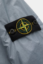Close-up side view of Stone Island Blouson 'Garment Dyed Crinkle Reps NY' rauchblau showing textured light blue microfiber fabric and black compass badge with yellow and green embroidery. Funktion trifft Style: Der sportive Blouson von STONE ISLAND besteh