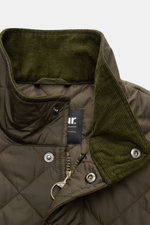 Nahaufnahme der olivgrünen Barbour Steppjacke 'Chelsea Sportsquilt' mit gestepptem Microfaserstoff, Stehkragen aus Cord und robustem Zwei-Wege-Reißverschluss, fotografiert aus schräger Perspektive.