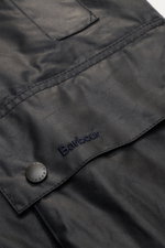 Close-up view of the Barbour Waxed jacket 'Beaufort' dark navy showing the waxed cotton fabric, flap pocket with snap button, and embroidered Barbour logo.

Description: A true classic in the British outdoor tradition: The waxed jacket 'Beaufort' by BARBO