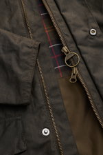 Nahaufnahme der Barbour Wachsjacke 'Sander' dark olive von vorne mit geöffnetem Reißverschluss, gewachste Baumwolle, kariertes Innenfutter, Cordkragen und großen Fronttaschen.