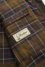 Nahaufnahme des karierten Innenfutters und der Innentasche der Barbour Wachsjacke 'Sander' dark olive mit Markenlabel.