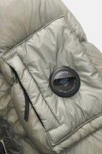 Close-up front view of the C.P. Company Down jacket 'DD Shell' sage, showing microfiber ripstop fabric, padded collar, and signature lens detail. For the lightweight down jacket 'DD Shell', C.P. COMPANY uses durable ripstop fabric and high-quality padding