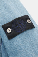 Close-up side view of the Stone Island Jeansjacke 'Indigo Denim-Rinsed' dunkelblau showing the black compass badge with blue embroidery and silver buttons on light blue denim fabric with typical cotton texture. Lässige Denim-Looks: Die aus indigogefärbtem