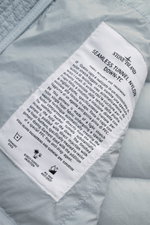 Close-up view of the Stone Island Daunenjacke 'Seamless Tunnel Nylon Down-TC' rauchblau label on light blue fabric, showing detailed care and fabric info. The jacket features high-quality microfiber, slight stretch, smooth touch, garment dyed, light duck 