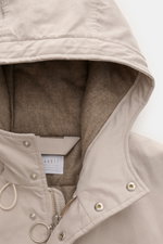 Close-up front view of the 04651/ A trip in a bag Parka 'MC26' sand, showing the hood and upper chest area with lightly waxed cotton exterior and soft wool-mix lining. Inspired by the Winter Olympics in Cortina and Milan, this parka features a high collar