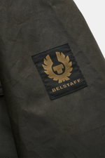 Close-up front-left sleeve view of the Belstaff Waxed Jacket 'Ground' dark olive, showing the BELSTAFF woven label on waxed cotton fabric.