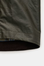 Close-up of the hem and cuff area of the Belstaff Waxed Jacket 'Ground' dark olive, photographed from the side showing waxed cotton, smooth feel, wind- and water-repellent fabric, adjustable sleeve cuffs, and checked lining fabric.