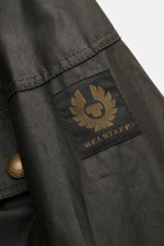 Nahaufnahme von Ärmeldetail mit BELSTAFF-Logo in gold auf schwarzem Patch der strapazierfähigen Belstaff Wachsjacke 'Trialmaster' dark olive, gewachste, wasserabweisende Baumwolle, robustem Reißverschluss und regulierbaren Ärmelabschlüssen.