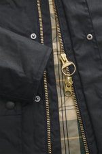 Close-up view of the Barbour Wachsjacke 'Reelin Wax' dark olive, showing waxed cotton fabric, gold-tone two-way zipper with ring pull, snap buttons, and classic tartan lining, highlighting its lightweight padding, slim fit, wind flap, cord collar, reinfor