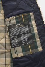 Close-up interior view of the Barbour Wachsjacke 'Reelin Wax' dark olive showing water-resistant waxed cotton, light padding, tartan lining, label-stitching, and care guide.